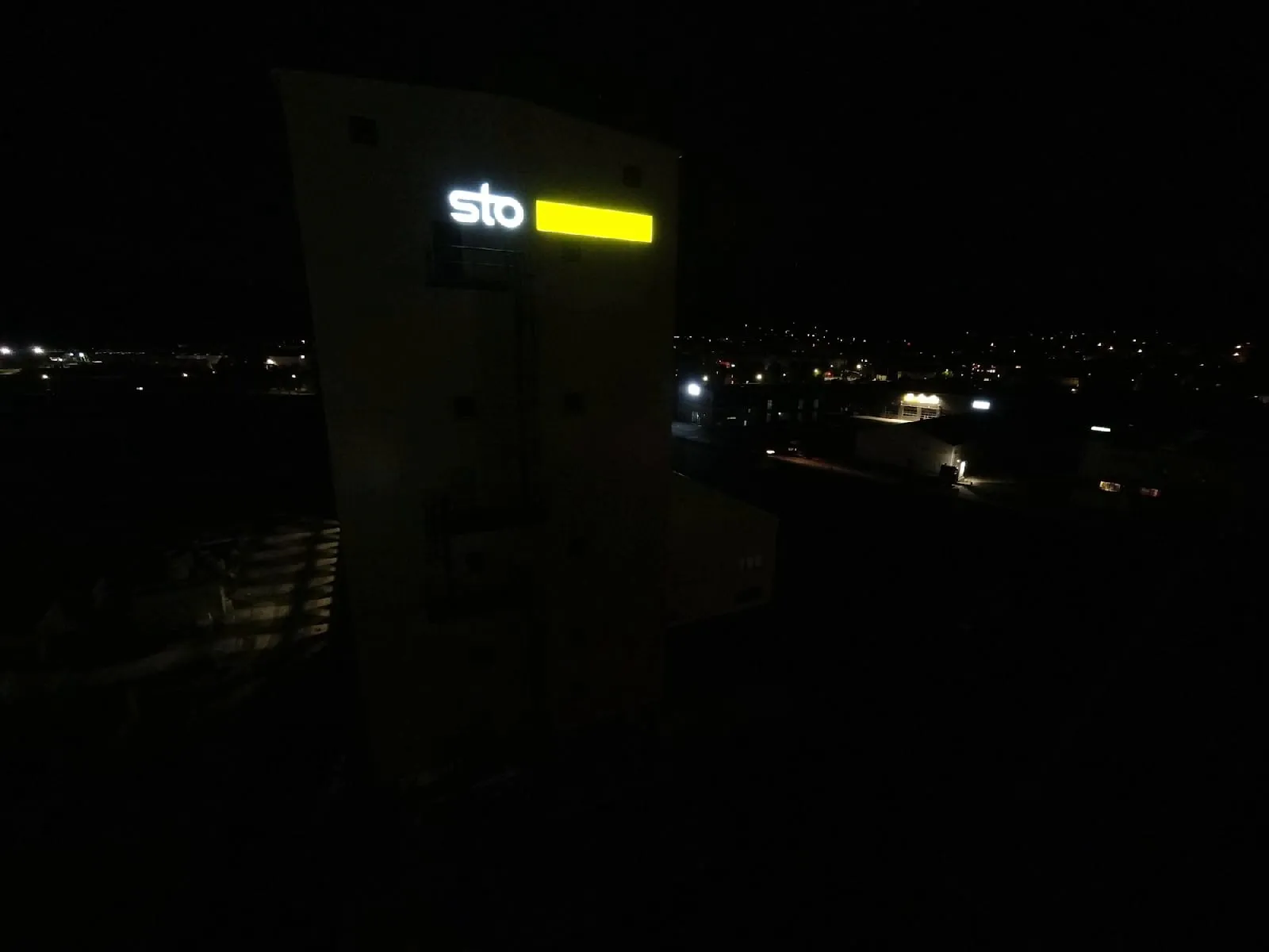 Derselbe Turm bei Nacht: Die Buchstaben „sto“ leuchten weiß, der Kasten daneben gelb