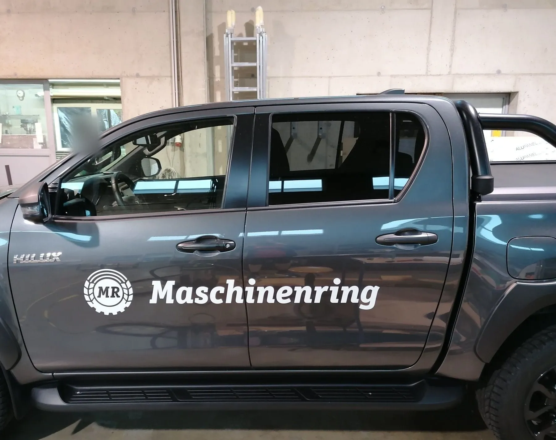 Grauer Toyota Hilux mit Maschinenring-Logo und Schriftzug an den Türen, Seitenansicht in unserer Werkstatt