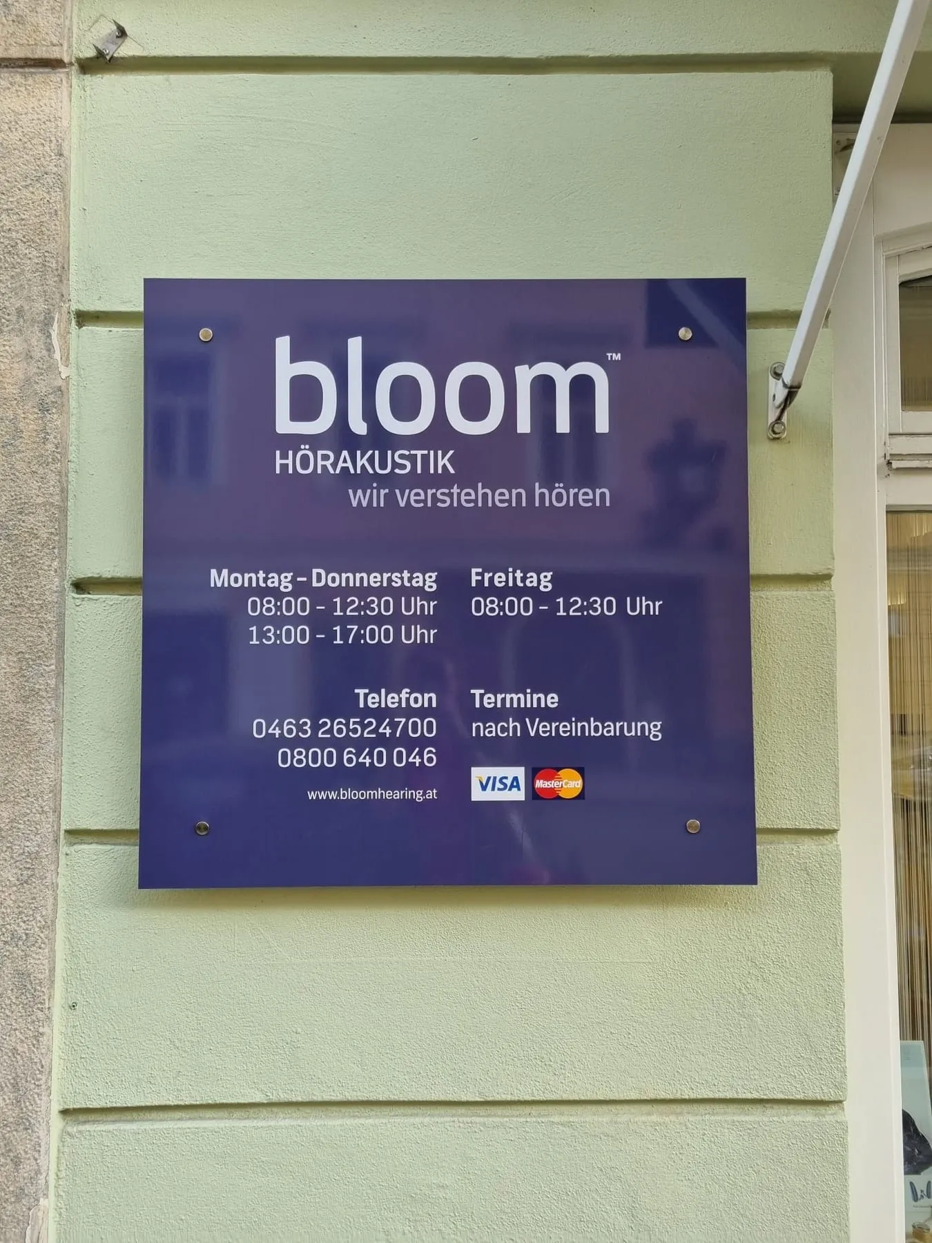 Dunkelblaues Schild mit Abstandhaltern an einer grünen Fassade, mit Logo und Öffnungszeiten