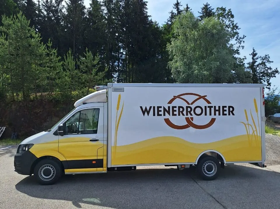 Transporter mit Kofferaufbau der Bäckerei Wienerroither, gelbe Welle und Ähren, Seitenansicht vor Wald