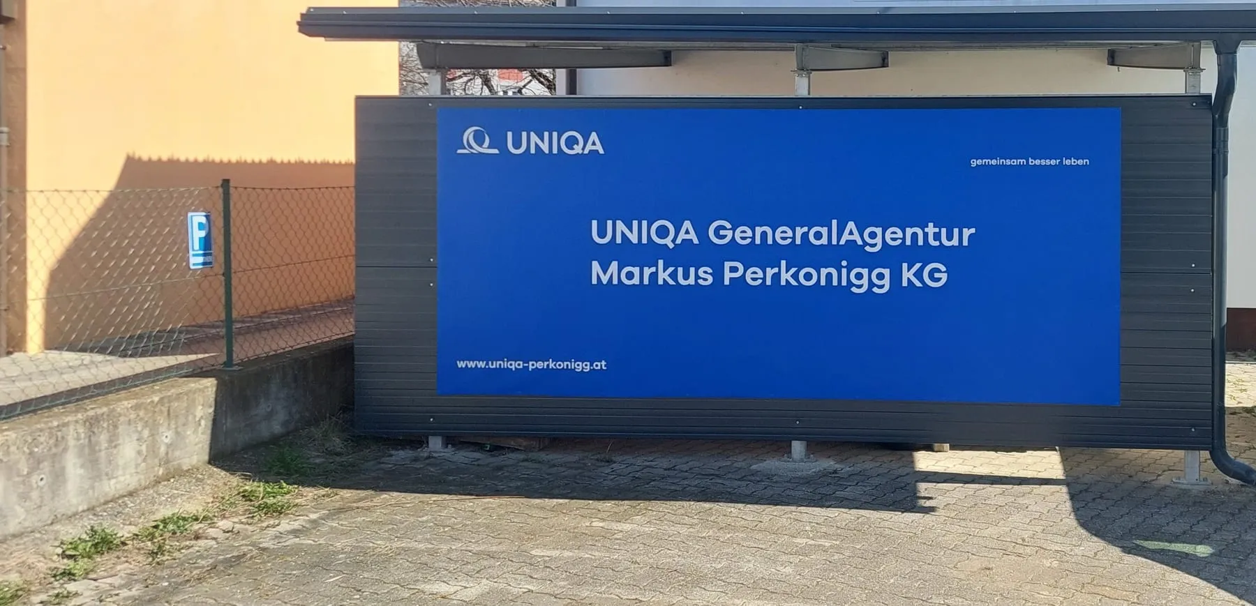 Große blaue Werbetafel einer UNIQA-Generalagentur unter einem Vordach