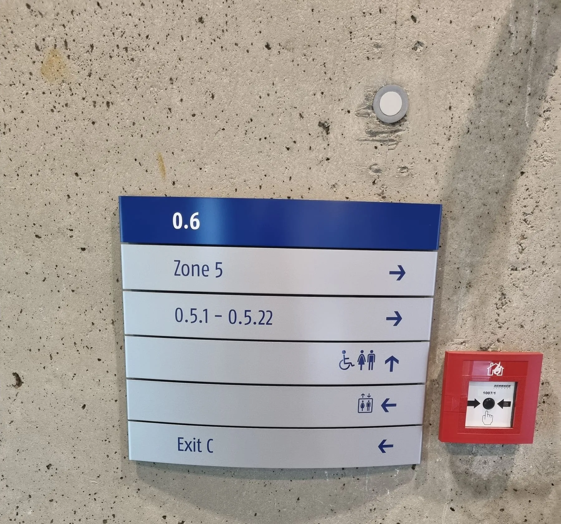 Wegweiserschild aus einzelnen Aluminiumzeilen mit Raumnummern, WC, Lift und Ausgang an einer Betonwand