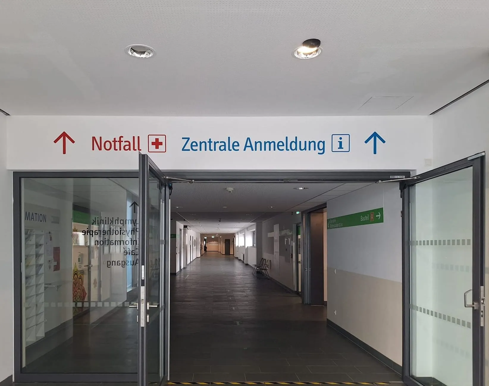 Klinik-Eingang mit Wandbeschriftung „Notfall“ und „Zentrale Anmeldung“ mit Pfeilen über offenen Glastüren