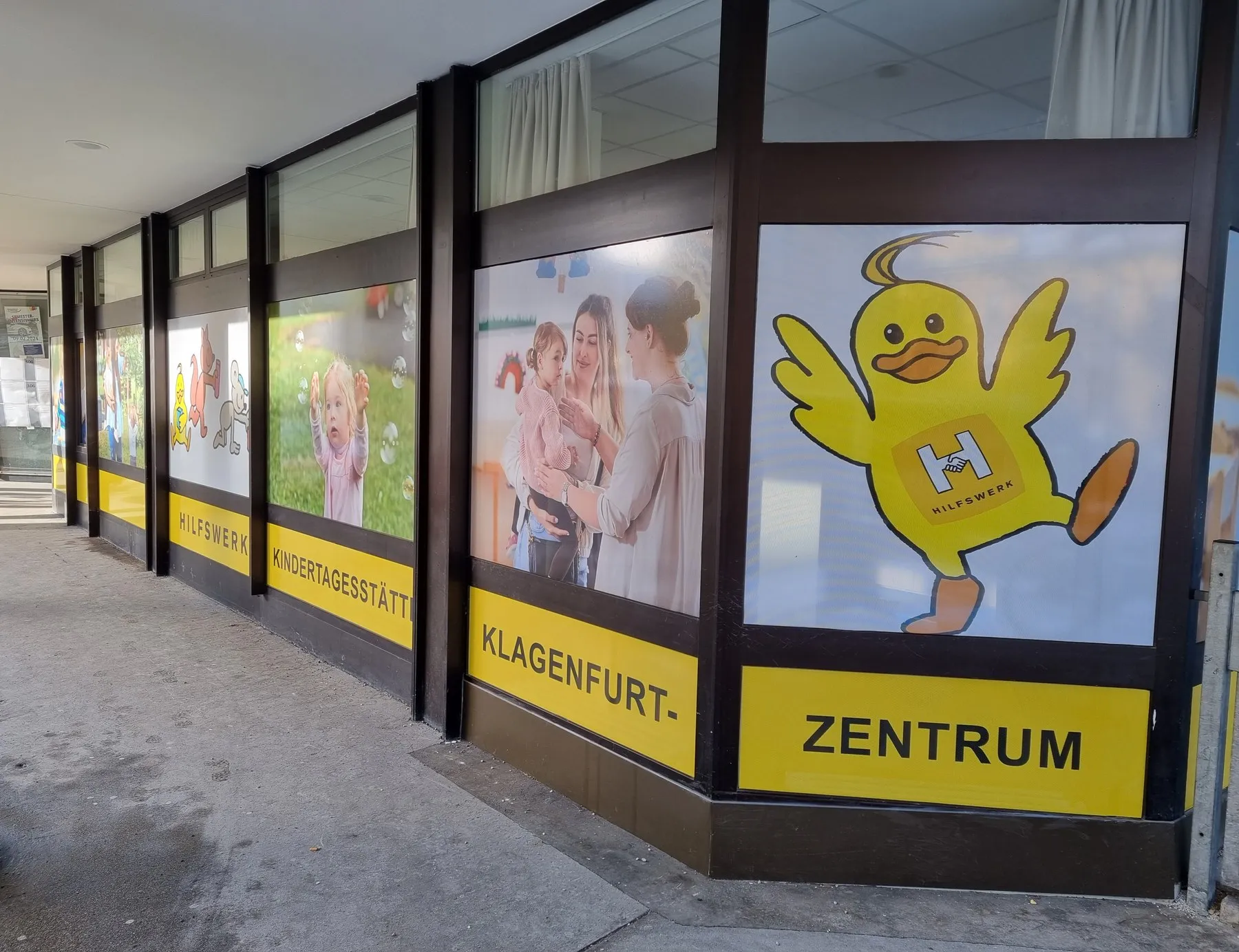Fensterfront der Hilfswerk-Kindertagesstätte Klagenfurt-Zentrum mit gelbem Band, Entchen und Fotomotiven