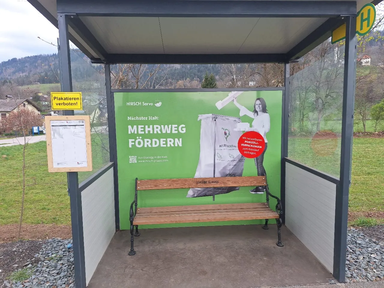 Buswartehäuschen mit bedruckter grüner Rückwand „Mehrweg fördern“ und Holzbank