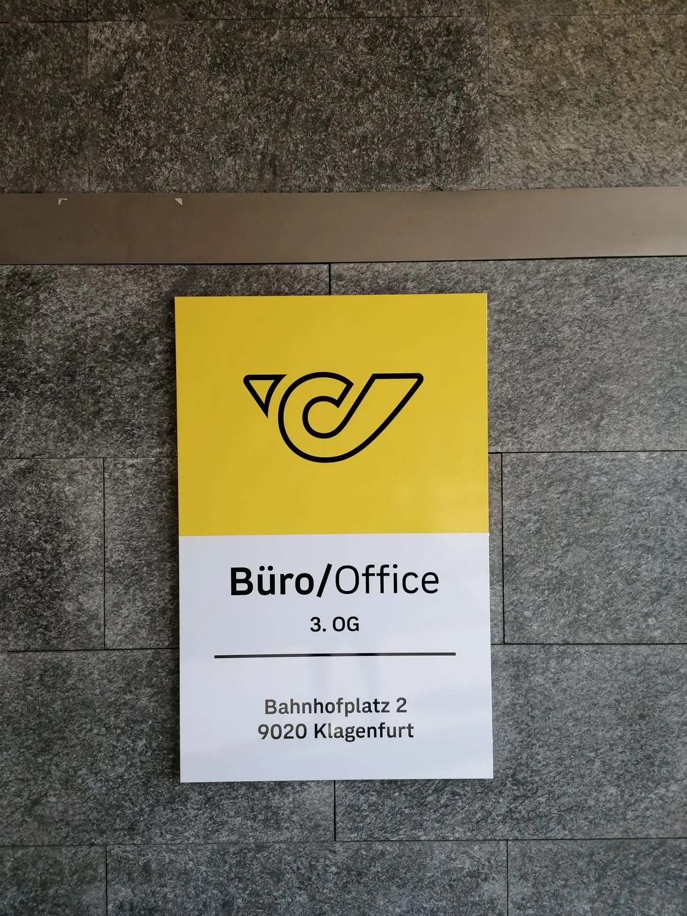 Gelb-weißes Wandschild mit Logo und Stockwerksangabe innen auf einer dunklen Steinwand