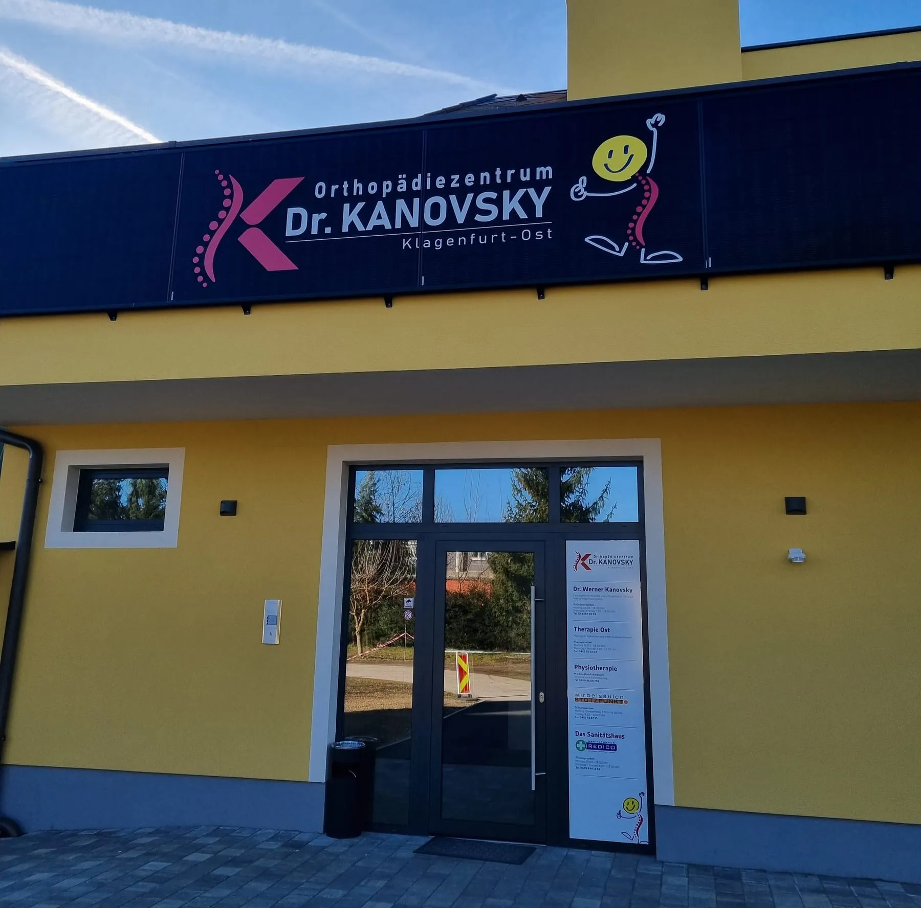 Dunkles Fassadenschild mit Logo und Schriftzug über dem Eingang eines gelben Gebäudes