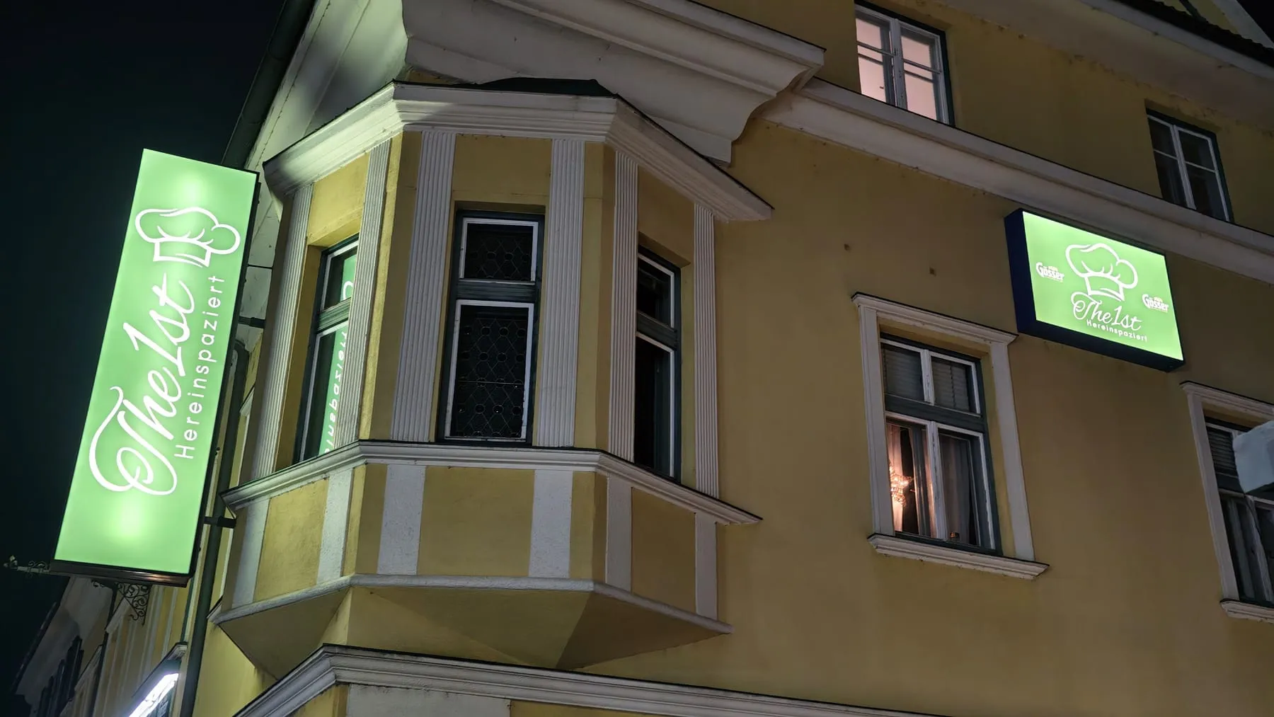Nasenschild und Leuchtkasten eines Restaurants leuchten grün an einer gelben Altbaufassade bei Nacht