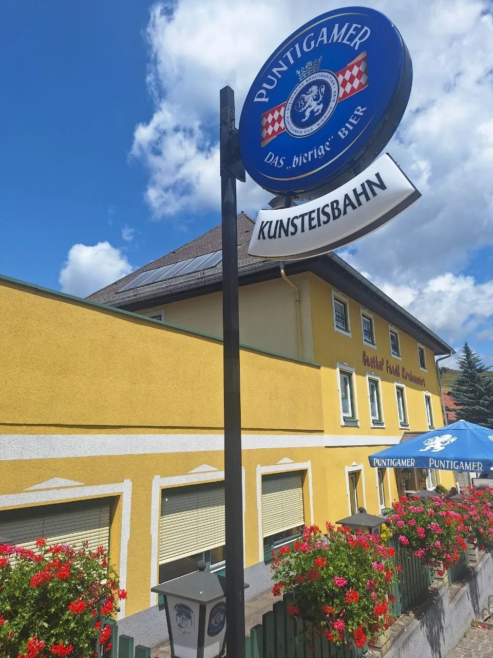 Runder Puntigamer-Ausleger mit Namensschild über dem Gastgarten eines gelben Gasthofs mit Blumen