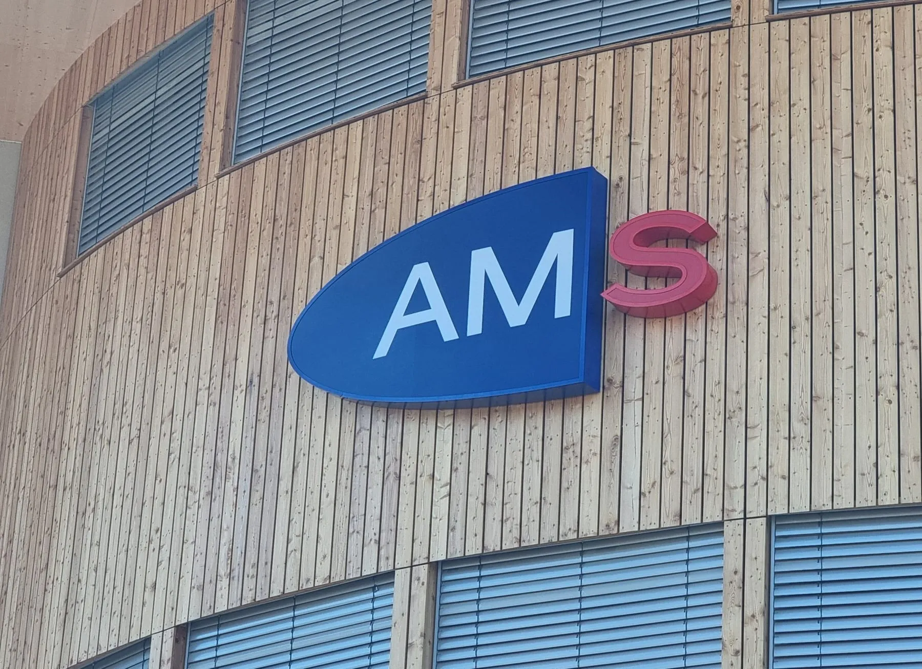 AMS-Logo als plastisches Element auf der runden Holzfassade des AMS Villach