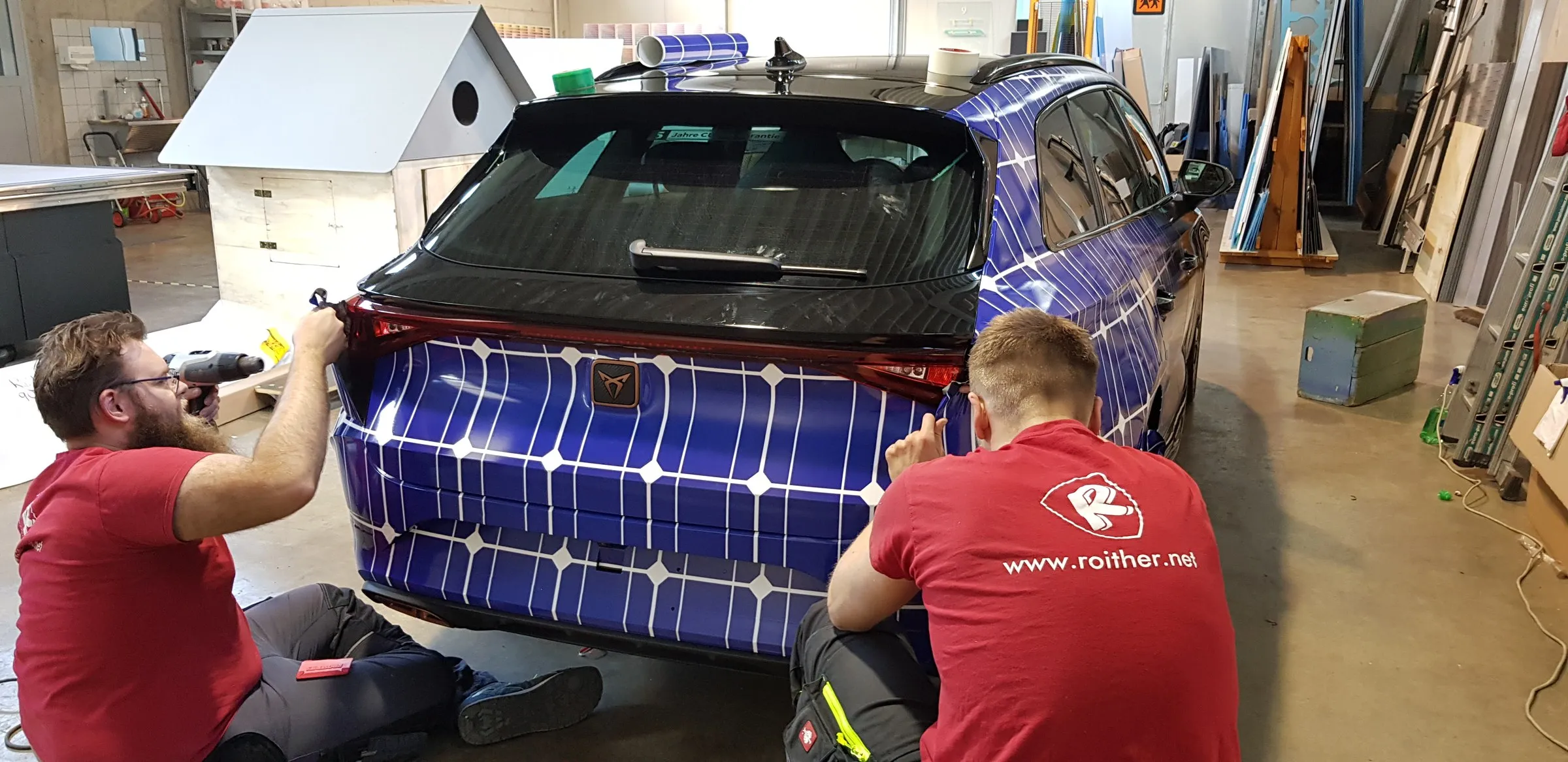 Zwei Monteure in roten Roither-Shirts folieren das Heck eines Autos mit Solarmotiv, in unserer Werkstatt