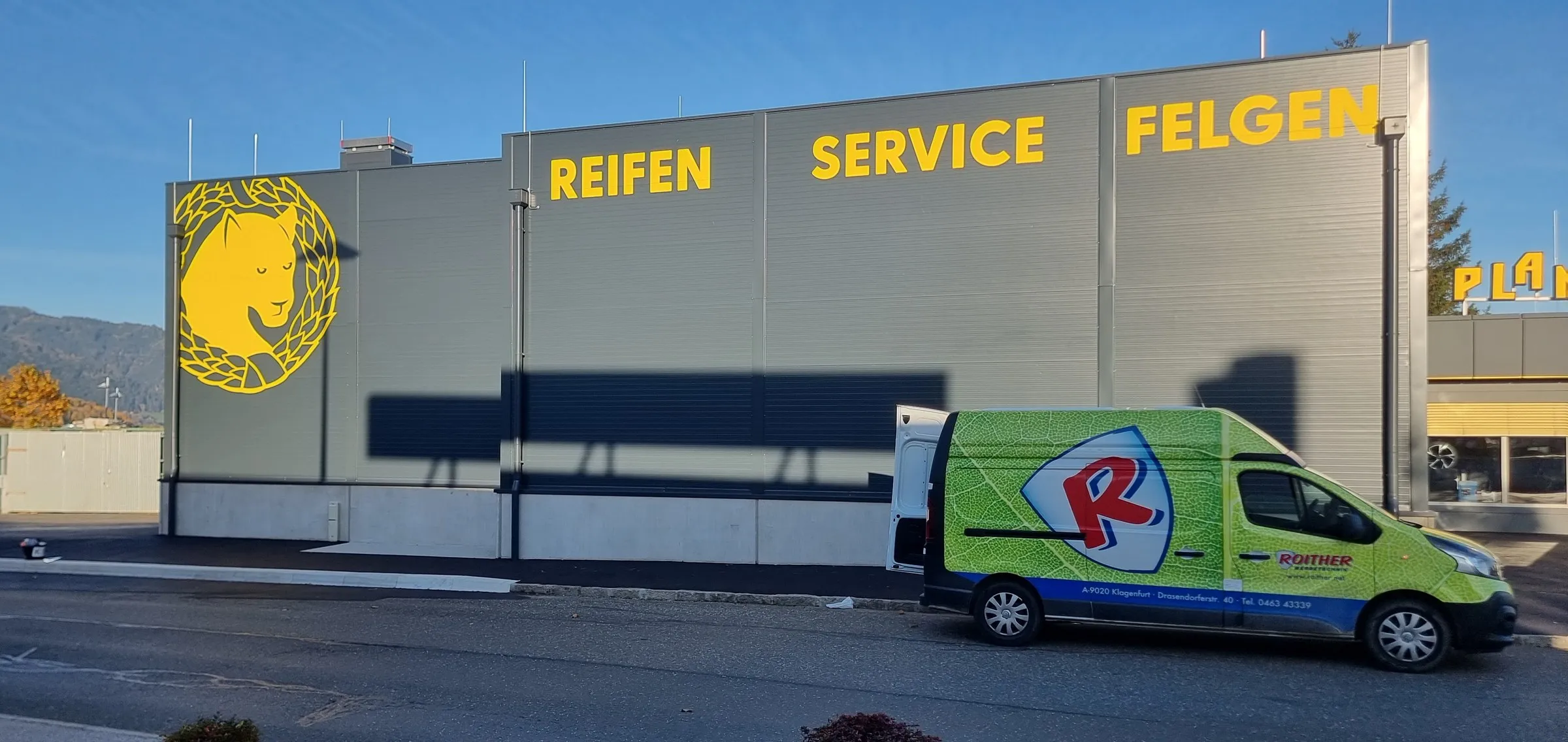 Gelbe Buchstaben REIFEN SERVICE FELGEN und Panther-Logo an der Halle von Plankenauer, davor unser Transporter